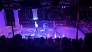 Performers in white costumes on an ice rink performing, with audience seated around and illuminated columns.
