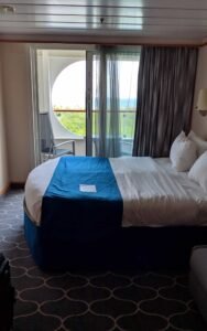 Hotel room with round bed, white and blue bedding, sliding door to balcony with chairs and ocean view.